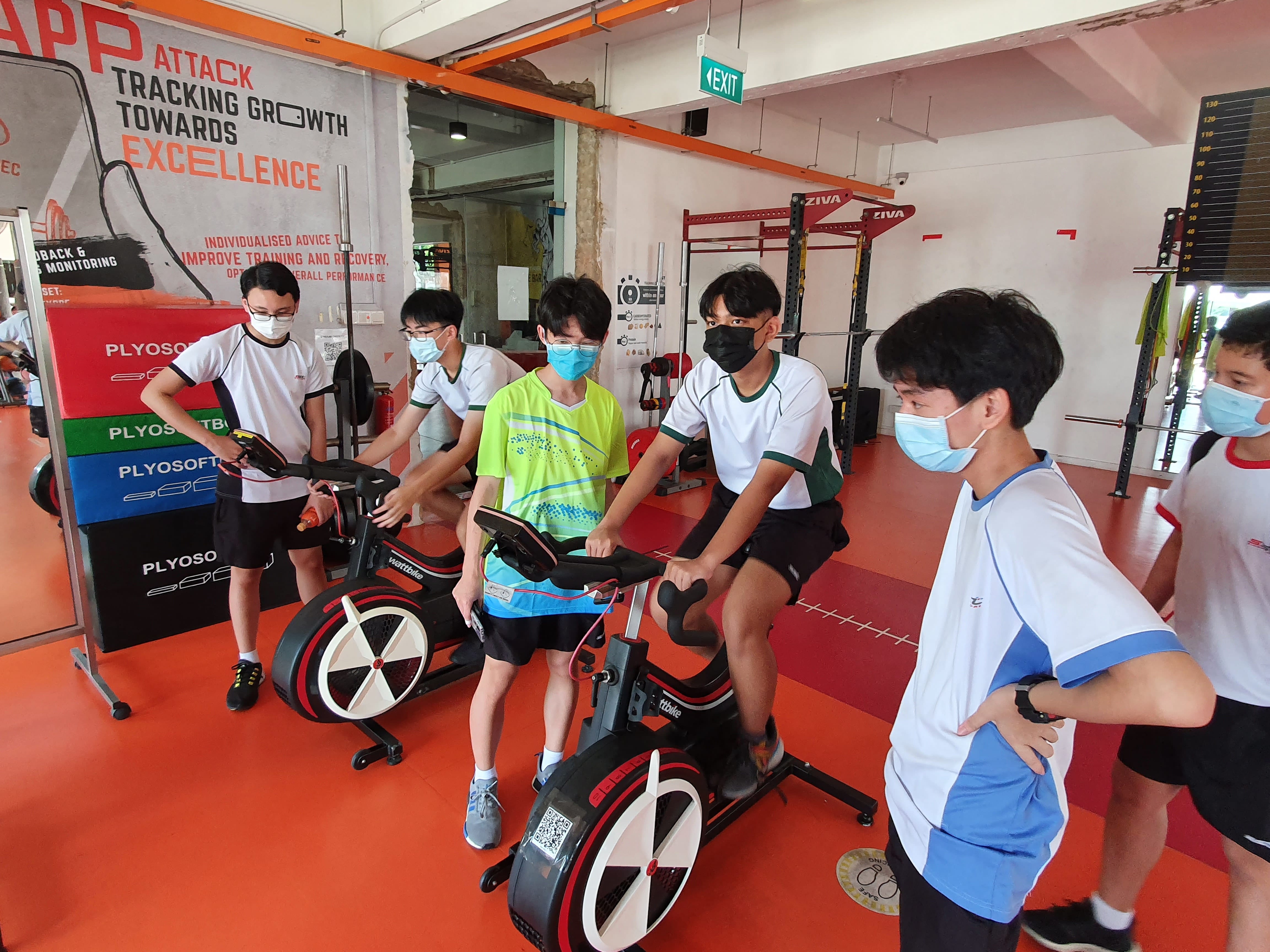 Several masked young students use stationary bikes in a gym with orange flooring and a motivational banner on the wall.